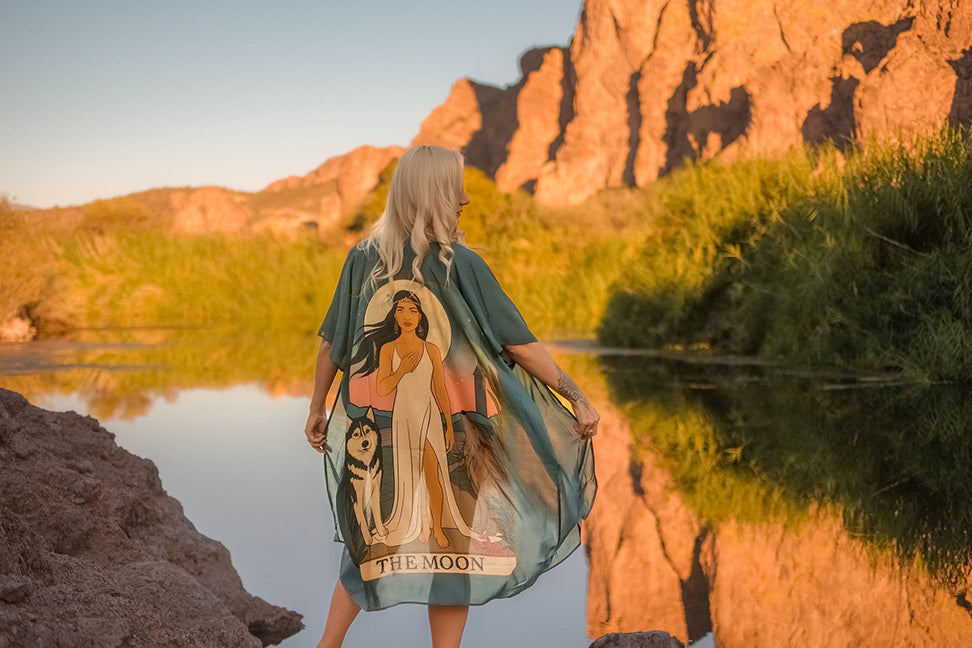 Marina Billinghurst wearing a dress with a Tarot design of a woman holding her heart and 'The Moon' text, standing by a reflective water body with rocky cliffs.
