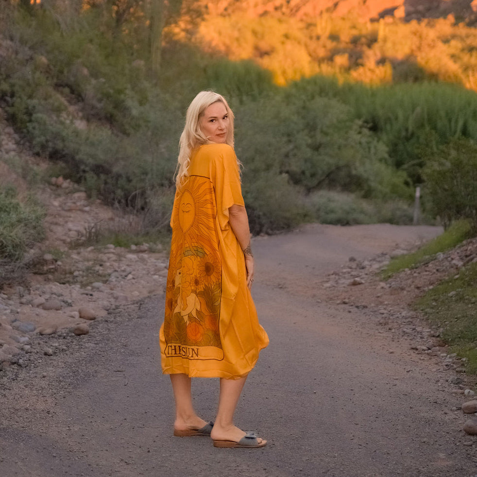 Marina Billinghurst in a yellow "Sun" Tarot Duster dress standing on a dirt path with desert landscape and cacti in the background.