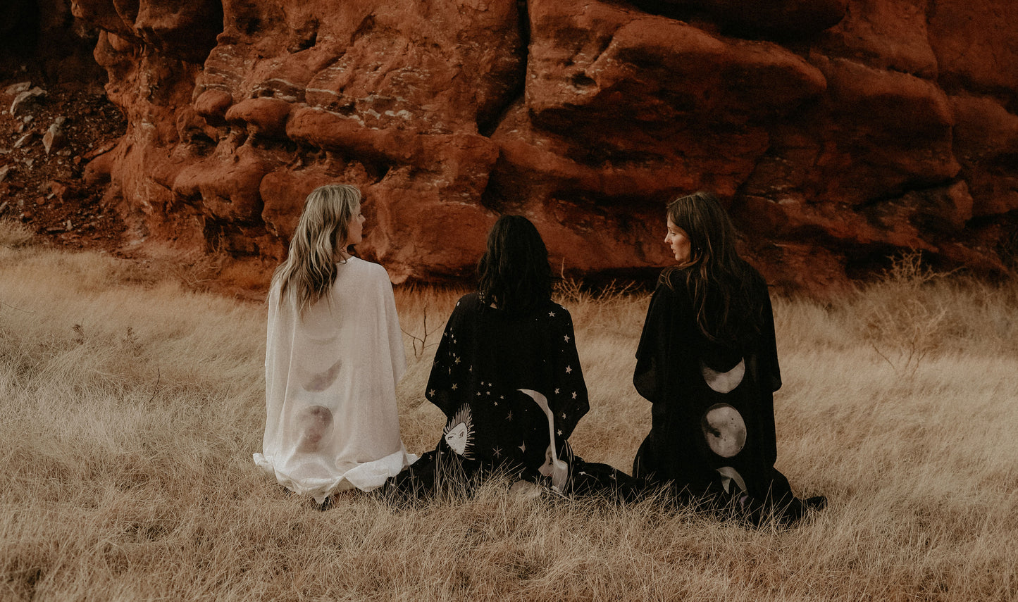 Three women sitting in a field with red rock formations in the background wearing art of marina kimonos with moon and moon phases art